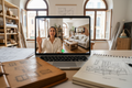 Laptop screen showing a video call between a Studio Collective designer in their Venice studio and a client showing their living room, with a layout sketch emerging in a notebook beside it